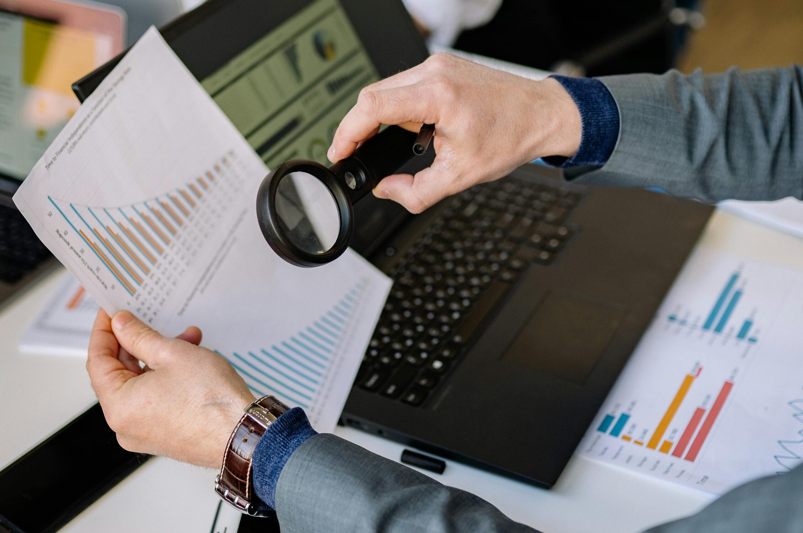 pexels-photo-7793173-7793173 Businessman examines data charts with a magnifying glass over a laptop in an office setting.
